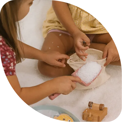 Two children sitting in a salt-covered room, using a pink scoop to fill a small canvas bag with white salt crystals.