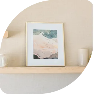 A framed piece of abstract coastal art sitting on a light wood floating shelf against a textured wall.
