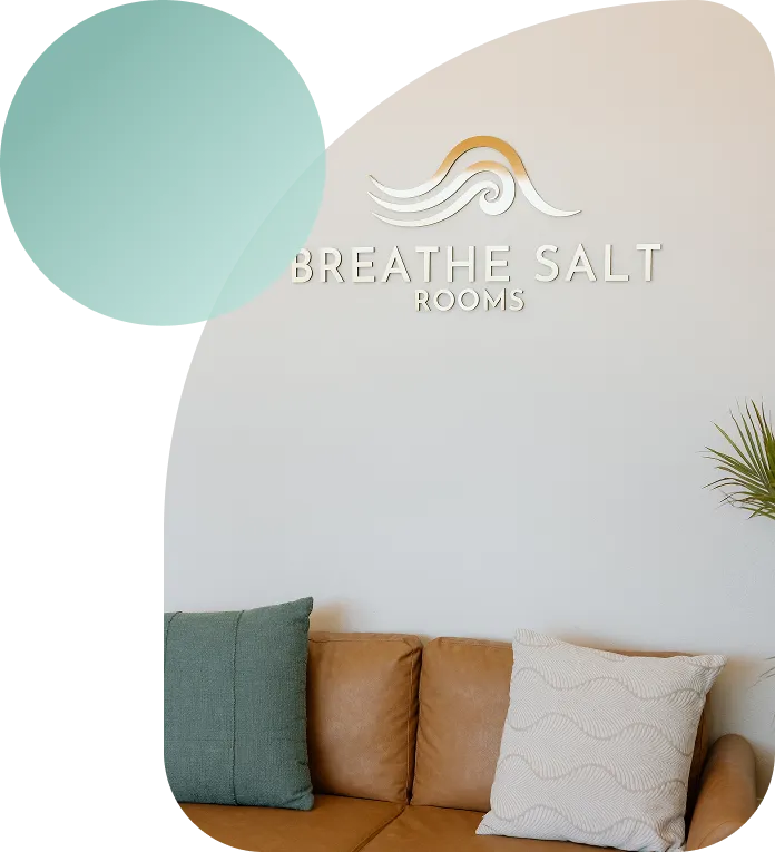 The welcoming reception area of Breathe Salt Rooms featuring a tan leather sofa, branded wall logo, and a large glowing Himalayan salt lamp.